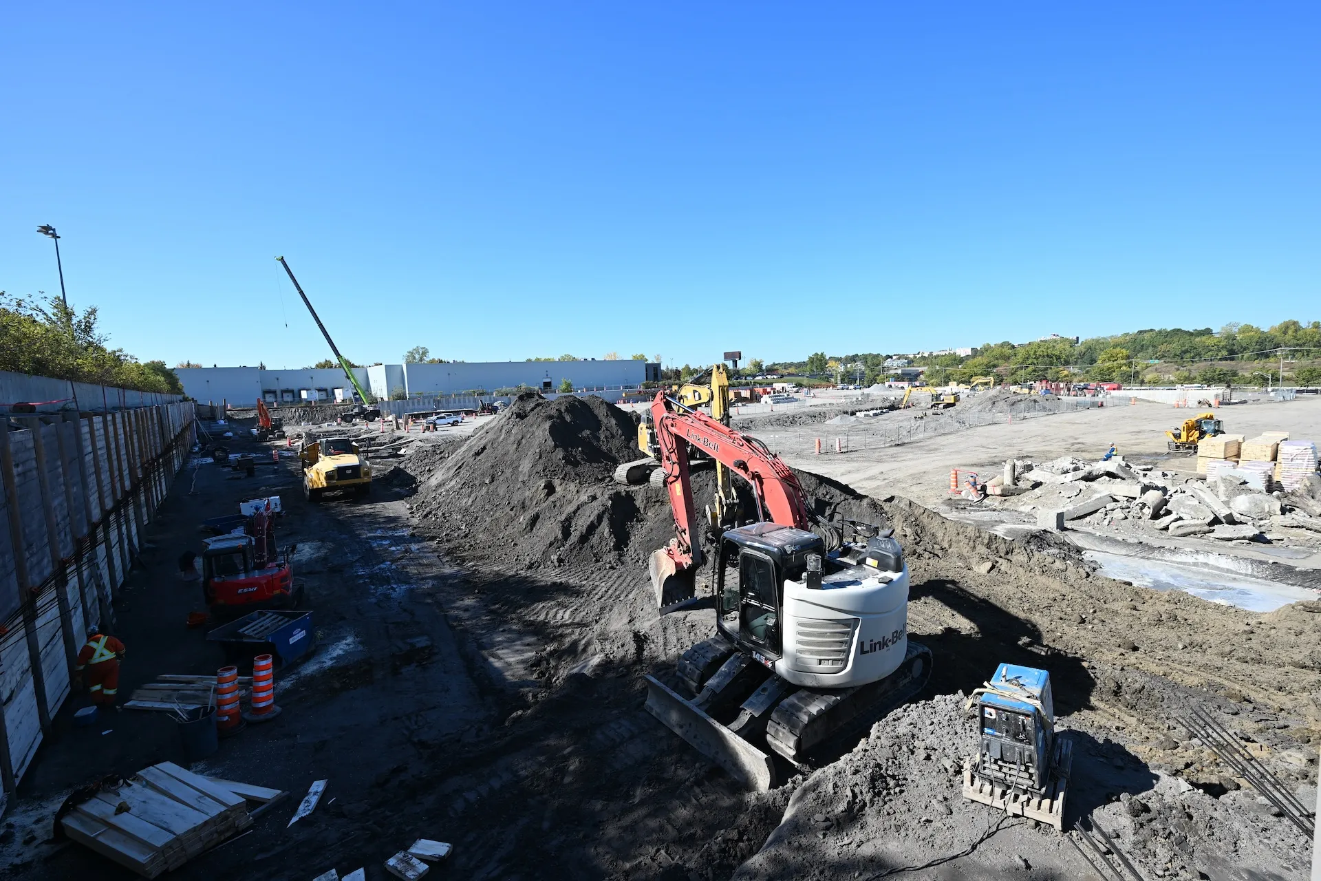 Construction site with heavy machinery, including excavators and a crane, working on large piles of dirt under a clear blue sky.