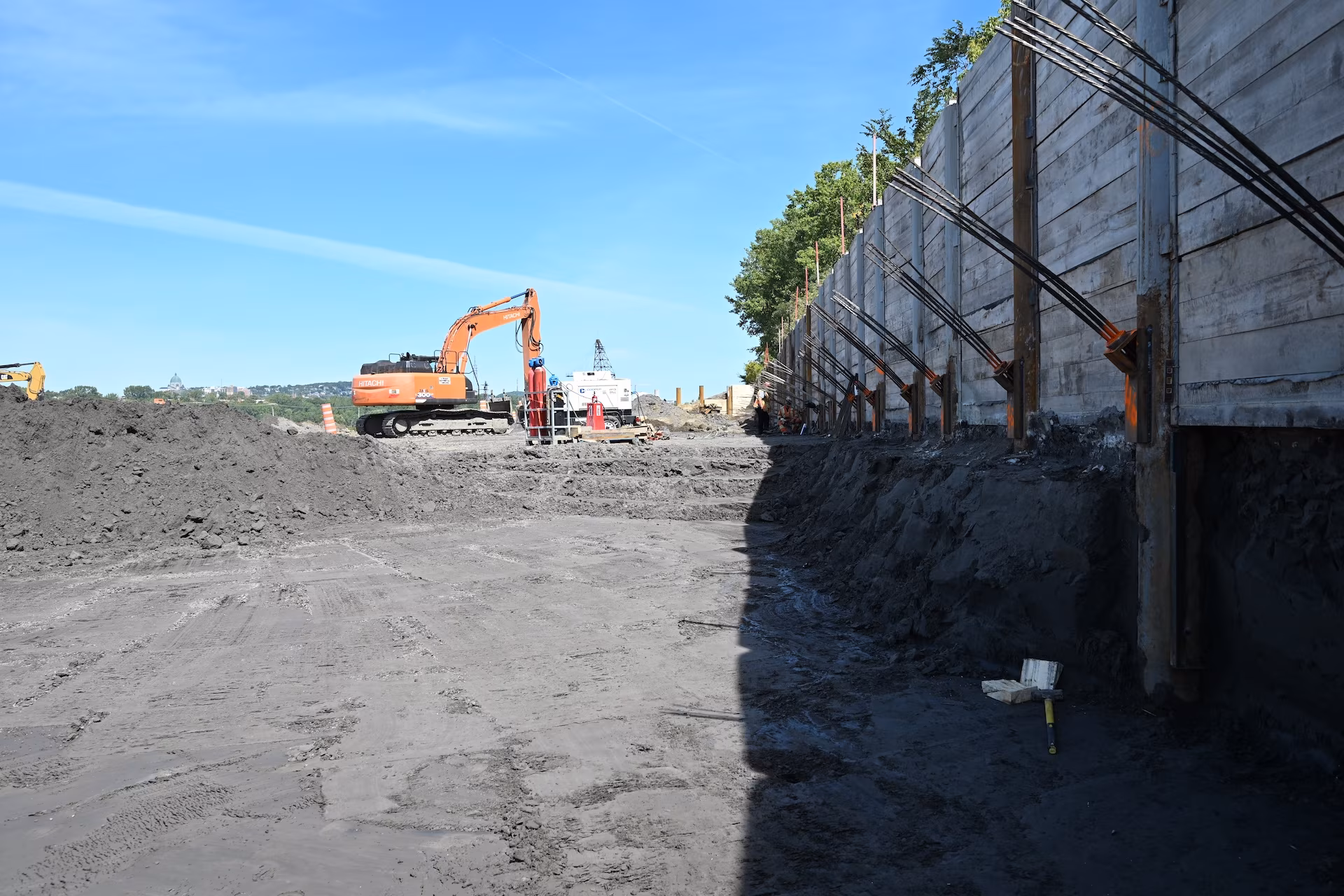 Construction site with an orange excavator and tall retaining wall supported by metal braces on a clear day.