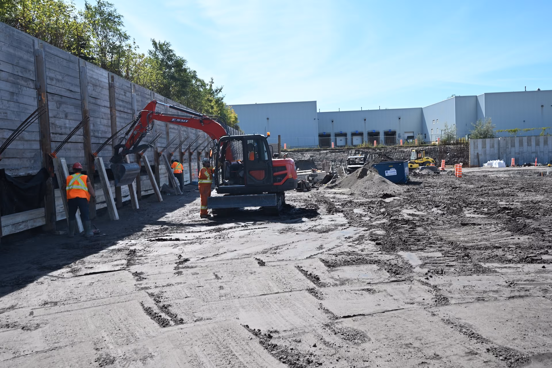 Construction site with workers in orange safety gear operating an excavator near a retaining wall with wooden supports and a warehouse building in the background.