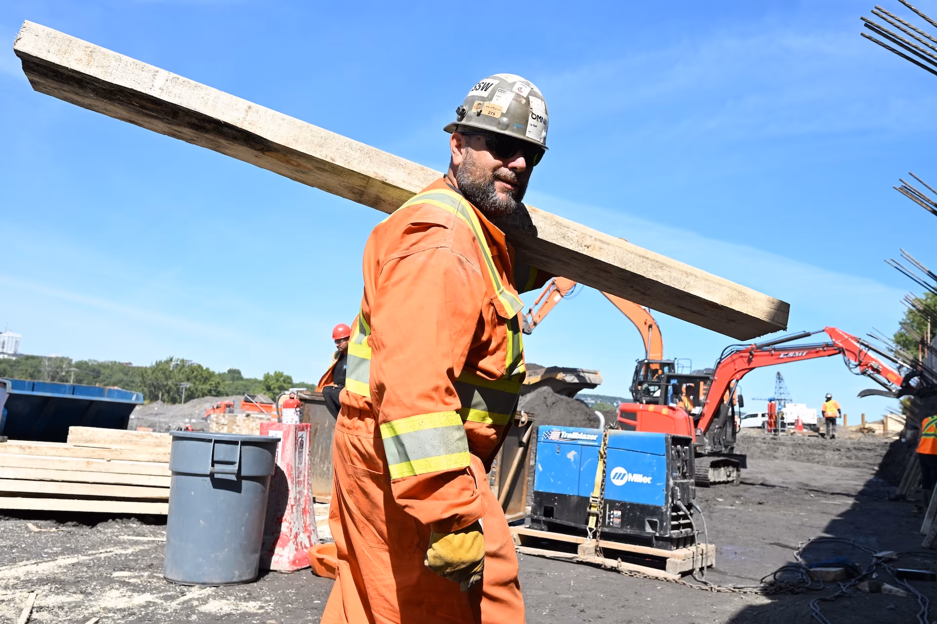 Construction worker in orange safety gear and helmet carrying a wooden plank at a construction site with excavators in the background.