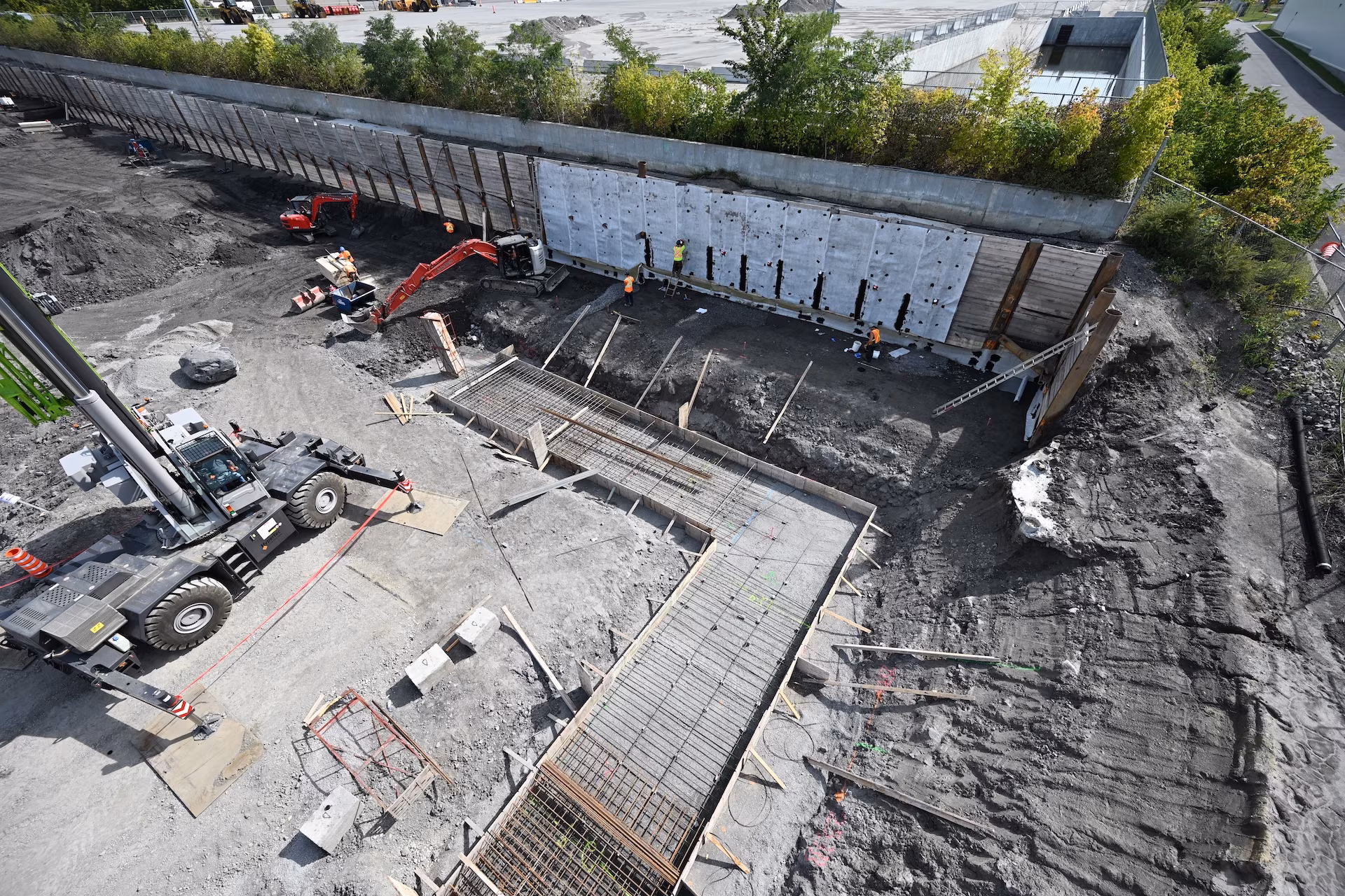Aerial view of a construction site with workers, excavation machinery, and rebar framework for foundation pouring beside a retaining wall.