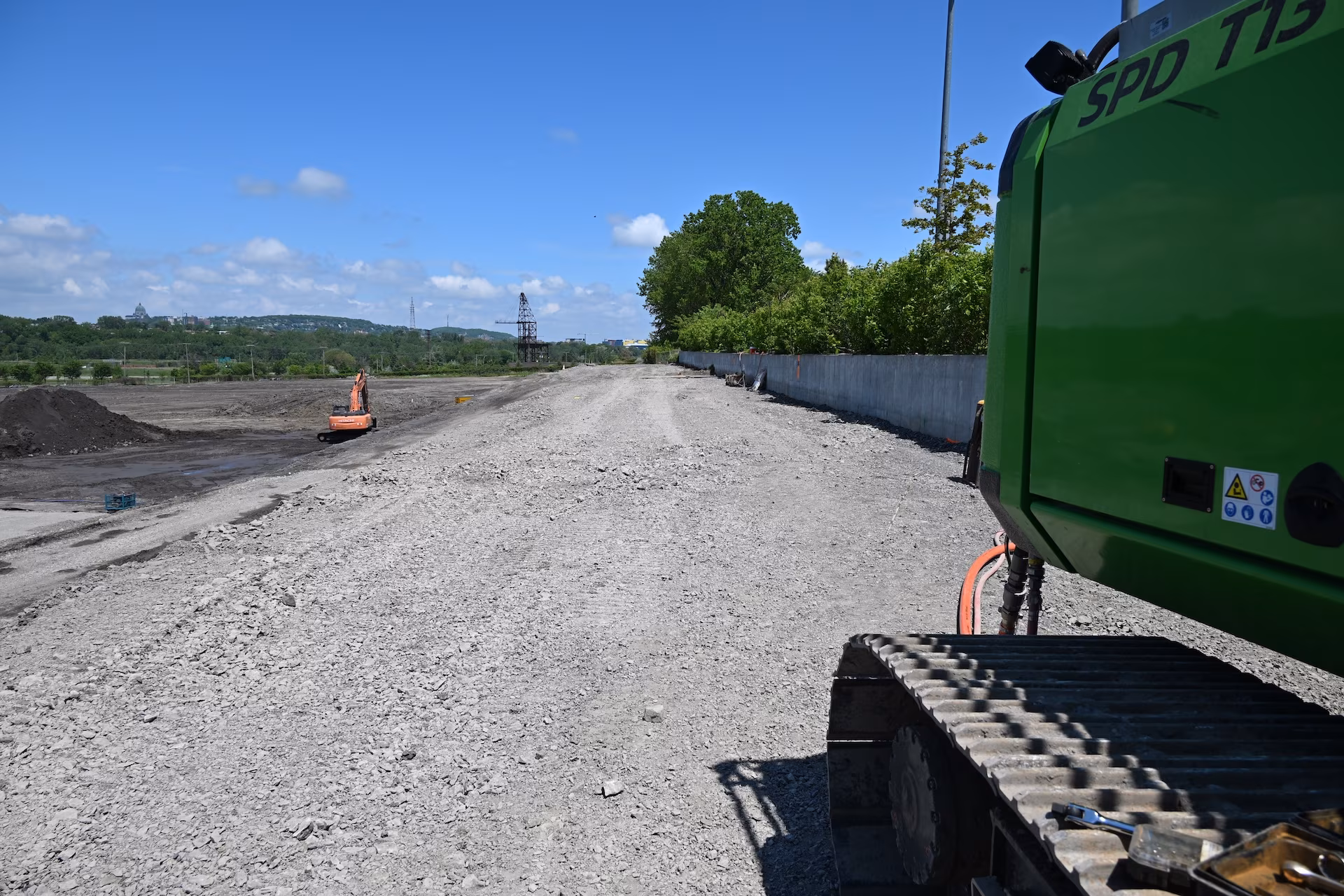 Construction site with gravel road, excavators, and a concrete wall under a blue sky.