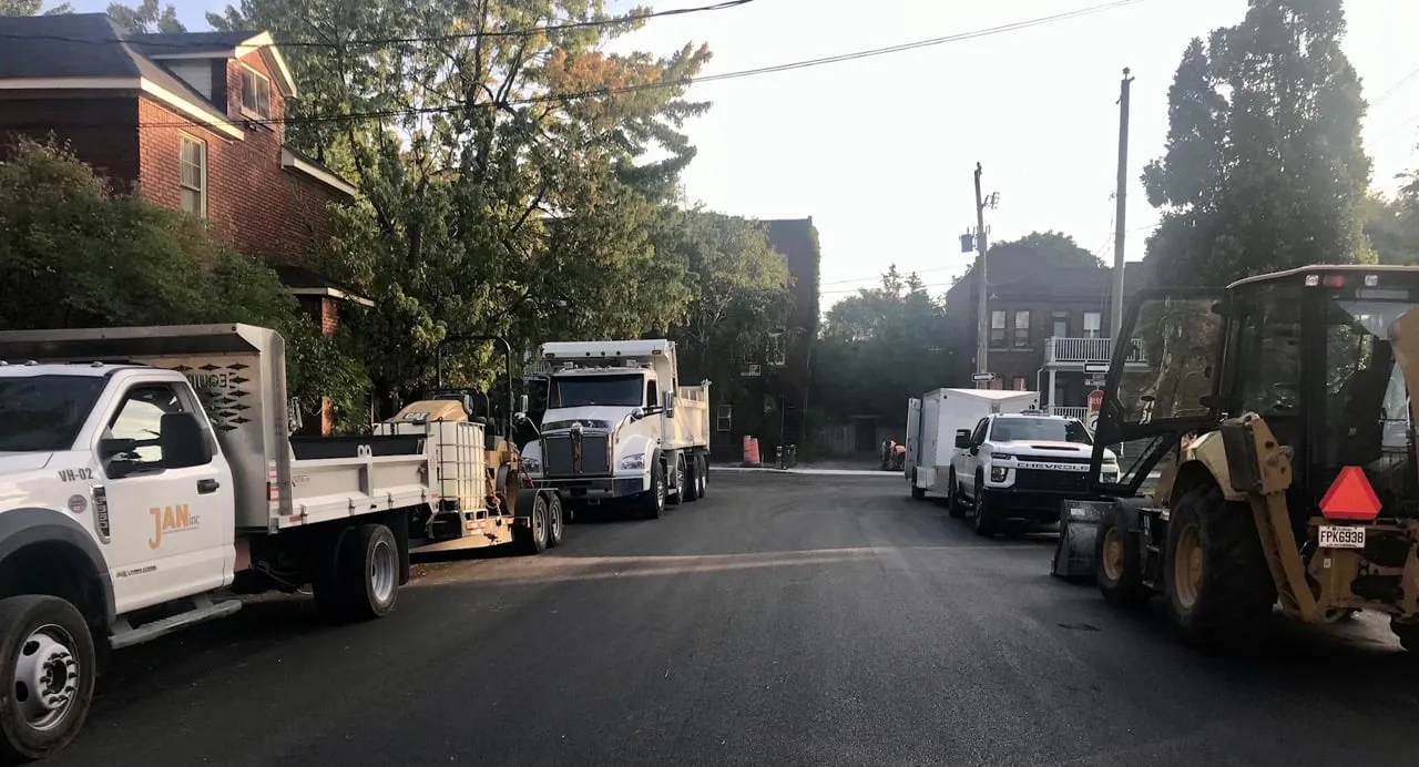Street view with several parked work trucks and construction equipment on both sides, bordered by trees and brick buildings.